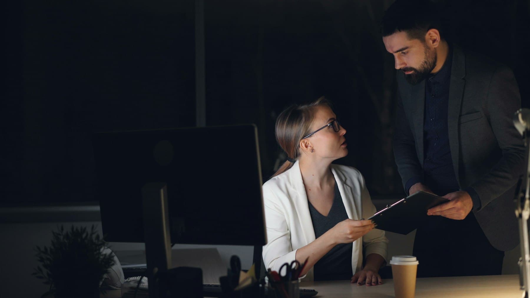 Dos profesionales revisando un expediente frente a un monitor en una oficina con luz tenue.